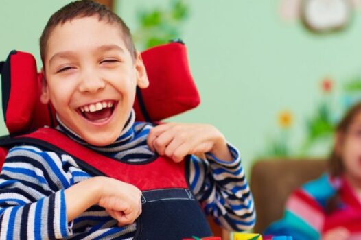 A cheerful child with short hair sits in a supportive wheelchair, smiling widely while wearing a striped shirt and red padding. In the softly blurred background, another child sits in a colorful classroom setting, suggesting a warm, inclusive learning environment.